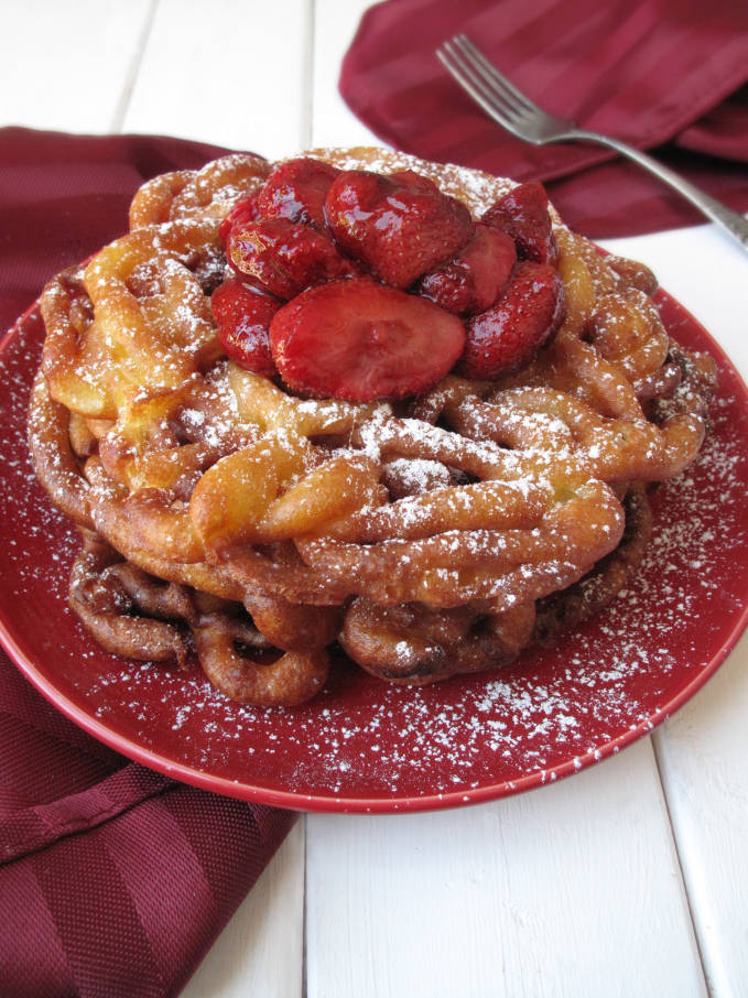 Fresh funnel cake dusted with icing sugar and served with warm strawberries, just like at the carnival! Click to grab the super simple and yummy recipe.