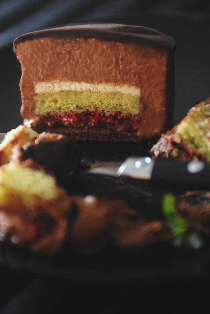 Ambroisie cake with biscuit joconde au chocolat, biscuit aux pistaches, homemade raspberry jam, dark chocolate mousse, pistachio mousse, and mirror glaze. Click to get the mouthwatering recipe!